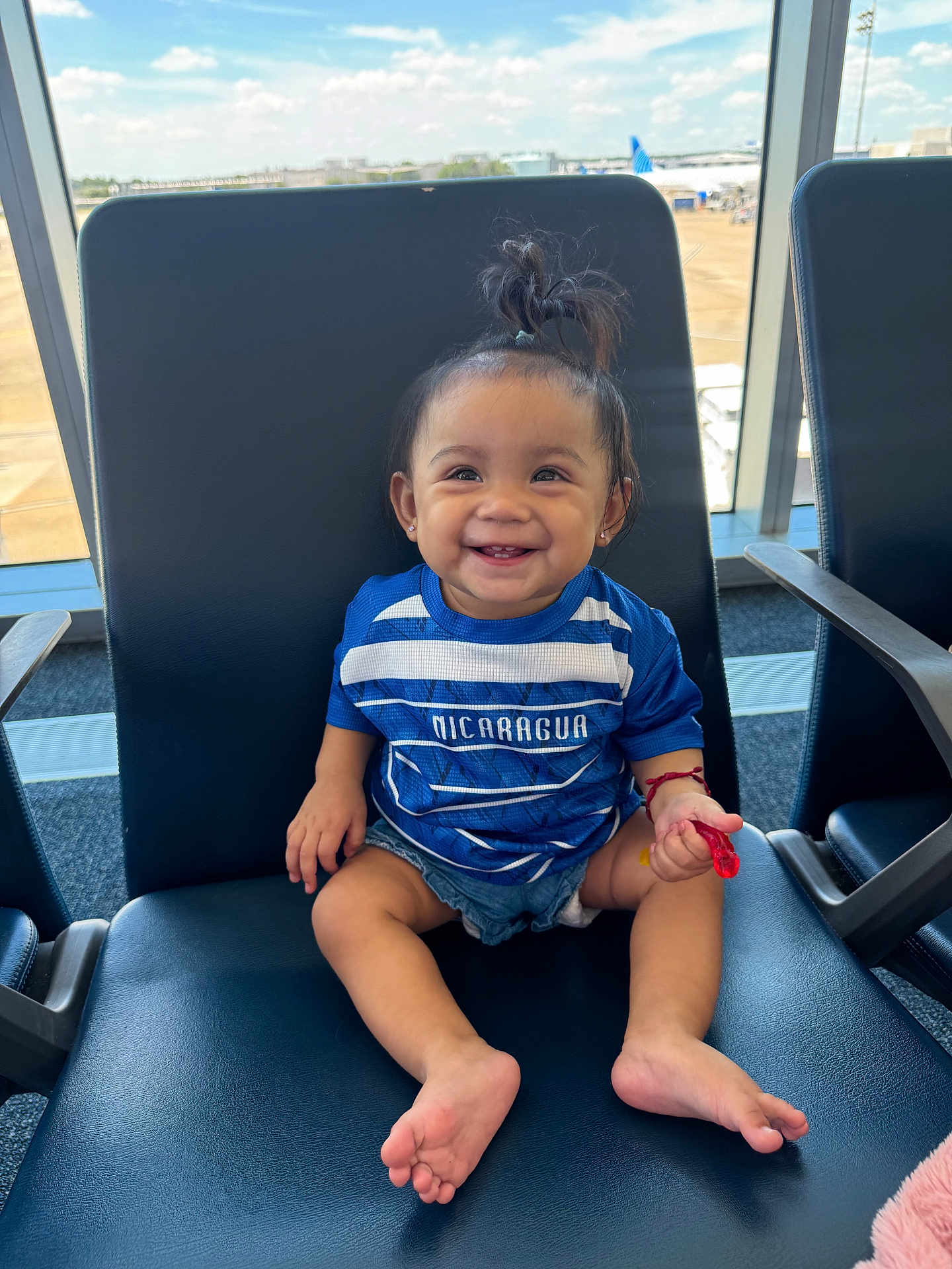 Sara Alina is registered to the contest to win money with this photo: toddler, child, smiling, sitting, airport, chair, blue_shirt, denim_shorts, pony_tail, happy, indoor, window, daylight, runway, airplane, footwear_free, person, cute, casual_clothing, waiting_area
