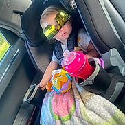 Astella is registered to the contest to win money with this photo: toddler, car_seat, sunglasses, blanket, toy, sippy_cup, child, car_interior, seatbelt, window, door, furniture, person, plastic, clothing, head, face, travel, baby, safety
