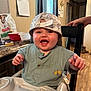 Parker is registered to the contest to win money with this photo: baby, high_chair, smiling, hat, green_shirt, kitchen, calendar, dog_food, countertop, window, curtains, hand, chair, floor, person, happy, indoor, tableware, food_container, home