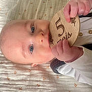 Fañch participe au concours pour gagner de l'argent avec cette photo : baby, infant, blue_eyes, hands, wooden_plaque, milestone, blanket, pattern, chewing, lying_down, cute, child, person, closeup, skin, face, white_clothing, soft_lighting, indoor, adorable