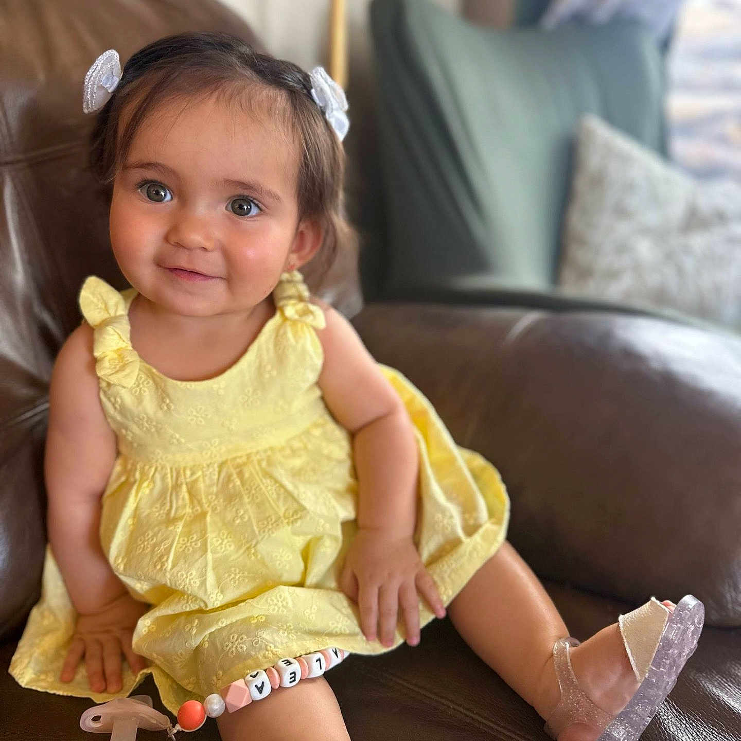Azalea is registered to the contest to win money with this photo: beaded_pacifier_clip, child, childhood, cute, eyes, face, footwear, hair_bows, happy, indoors, leather_couch, legs, pacifier, person, portrait, sitting, skin, smile, toddler, yellow_dress