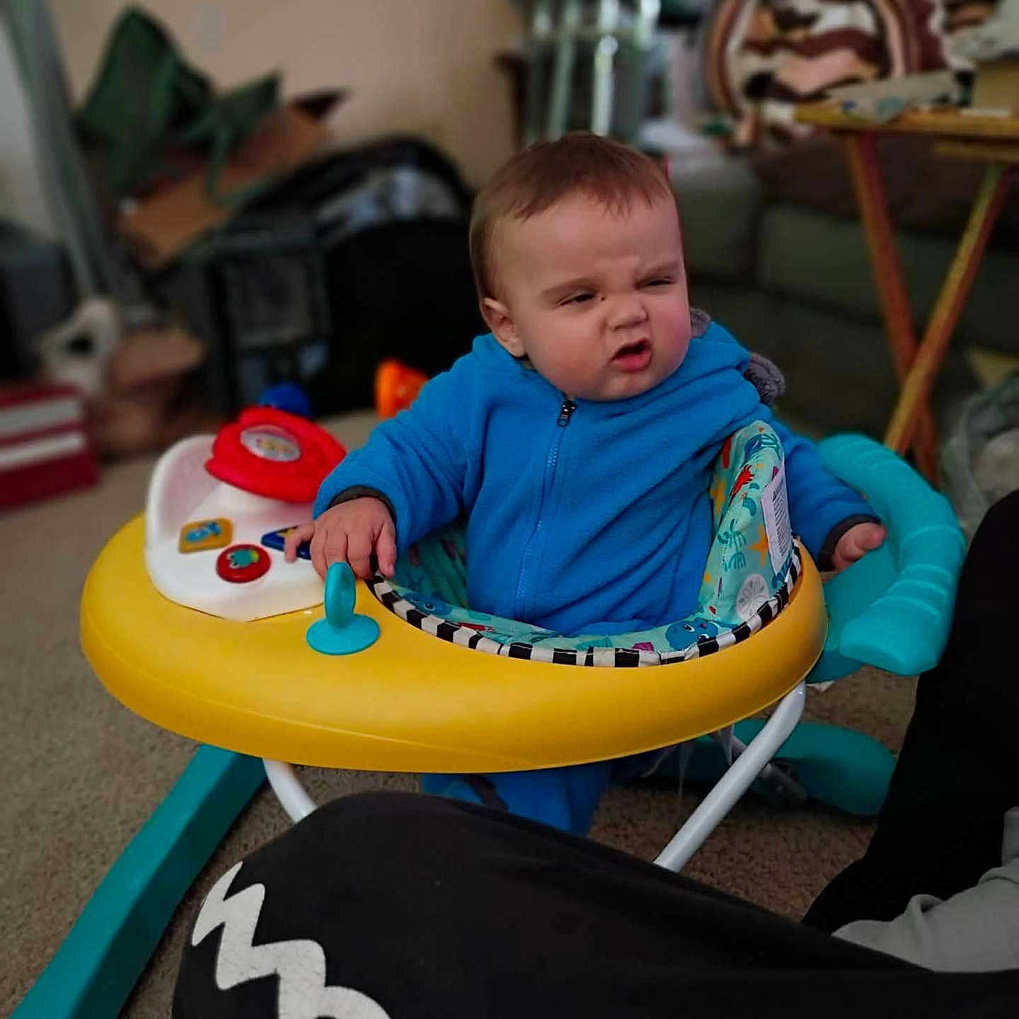 Killian is registered to the contest to win money with this photo: baby, baby_seat, blanket, blue_jumpsuit, candid, carpet, child, colorful, facial_expression, frowning, hand, infant, lamp, living_room, playtime, sofa, table, toddler, toy, walker