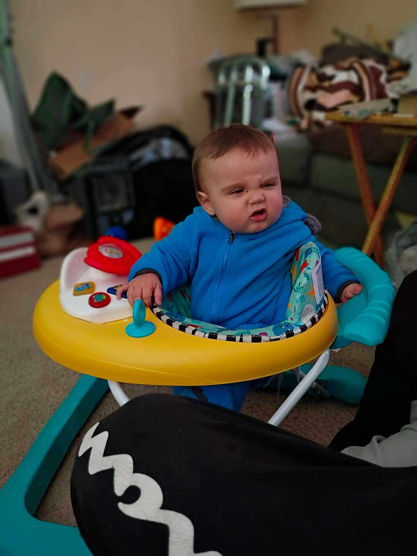 Killian is registered to the contest to win money with this photo: baby, infant, child, toddler, walker, toy, blue_jumpsuit, frowning, facial_expression, living_room, sofa, blanket, carpet, playtime, baby_seat, hand, colorful, lamp, table, candid