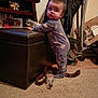 child, baby, toddler, pajamas, bear_prints, standing, ottoman, carpet, bottle, living_room, furniture, jacket, coat_rack, photo_frame, family_photo, floor, belt, cardboard_box, shelf, candid