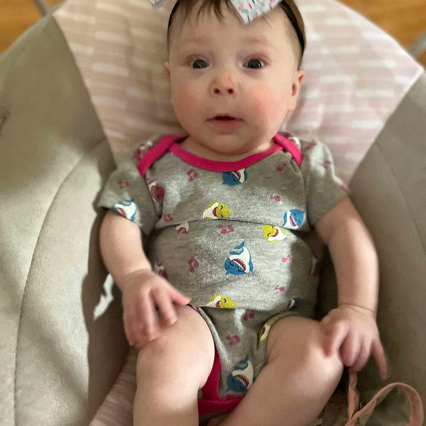 Cassidy is registered to the contest to win money with this photo: arms, baby, baby_seat, bow, child, clothing, cushion, cute, expression, gray, headband, indoor, infant, legs, onesie, person, pink, seat, shark_pattern, wide_eyes