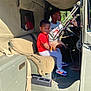 child, children, vehicle, car, military_vehicle, reflection, red_shirt, gray_pants, sneakers, steering_wheel, seat, window, sunlight, outdoor, daytime, person, portrait, interior, casual_clothing, curious