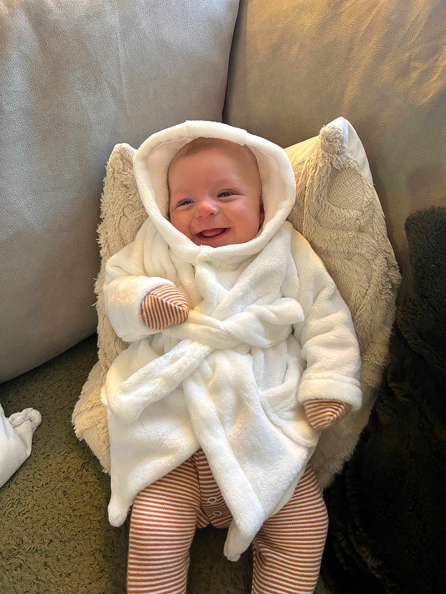 Arlo joined the competition — help win amazing prizes! baby, smiling, hood, robe, mittens, striped_clothing, pillow, cushion, carpet, indoor, person, infant, happy, cozy, soft_texture, clothing, portrait, child, cute, relaxed