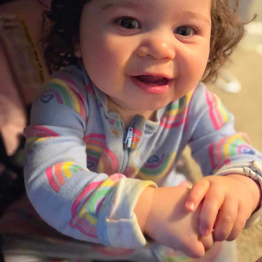Amelia is registered to the contest to win money with this photo: baby, child, curly_hair, smiling, onesie, rainbow_pattern, indoor, holding_hand, happy, cute, portrait, infant, top_knot, person, young_child, playful, warm_lighting, closeup, seated, family