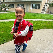 Otis is registered to the contest to win money with this photo: toddler, child, smiling, red_backpack, red_jacket, black_shirt, checkered_pants, blue_crocs, outdoor, grass, concrete_path, apartment_building, happy, person, walking, casual_clothing, young_child, playful, daylight, urban
