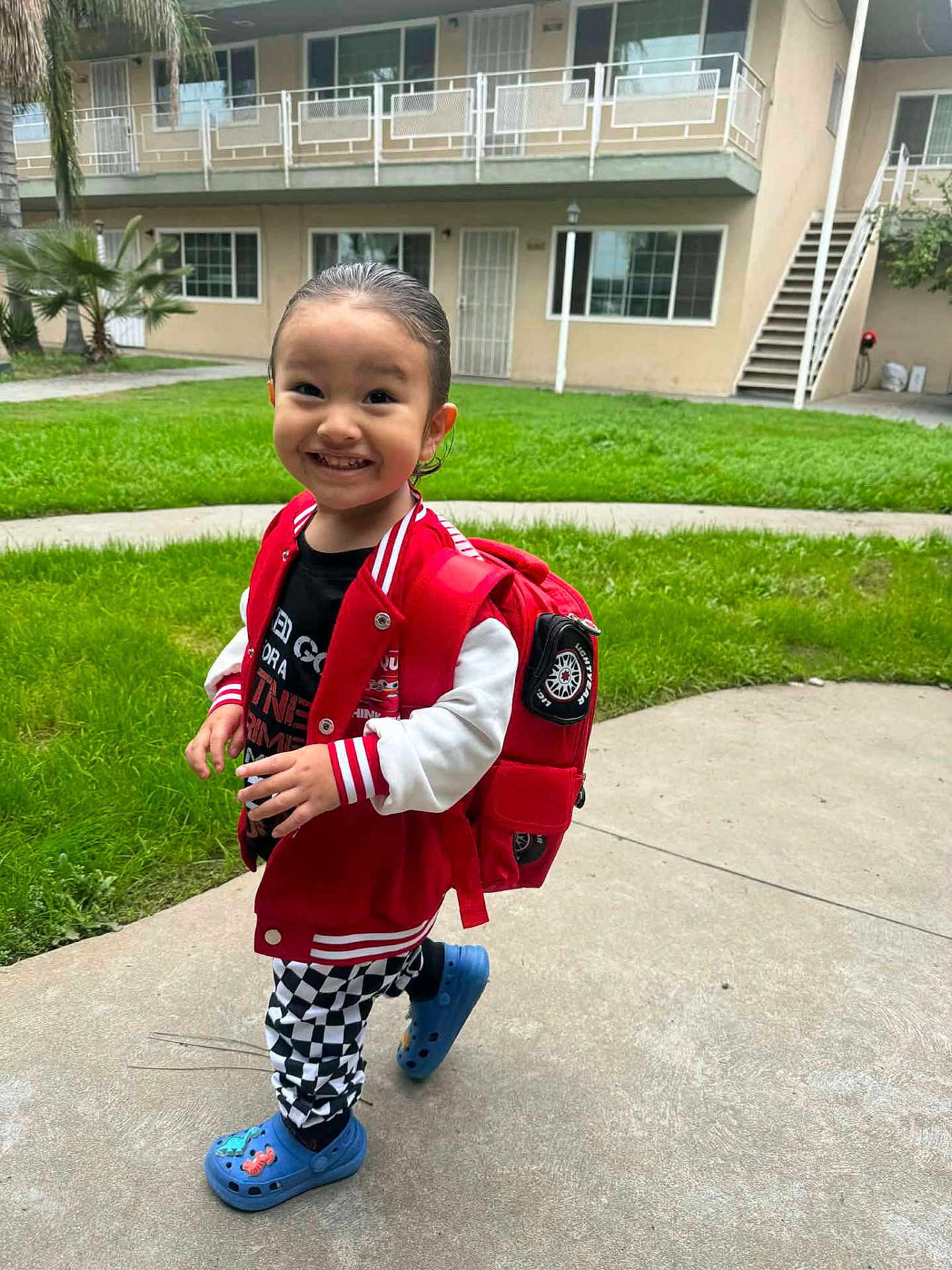 Otis is registered to the contest to win money with this photo: toddler, child, smiling, red_backpack, red_jacket, black_shirt, checkered_pants, blue_crocs, outdoor, grass, concrete_path, apartment_building, happy, person, walking, casual_clothing, young_child, playful, daylight, urban