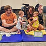 Spring is registered to the contest to win money with this photo: baby, child, adult, woman, man, indoor, floor, mats, stroller, blanket, yellow_clothing, glasses, carpet, happy, smile, play, family, people, interaction, seated