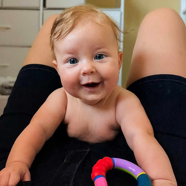 Spring is registered to the contest to win money with this photo: baby, blue_eyes, child, closeup, clothing, cute, expression, face, hands, happy, human, indoor, infant, joy, legs, person, playful, skin, smiling, toy