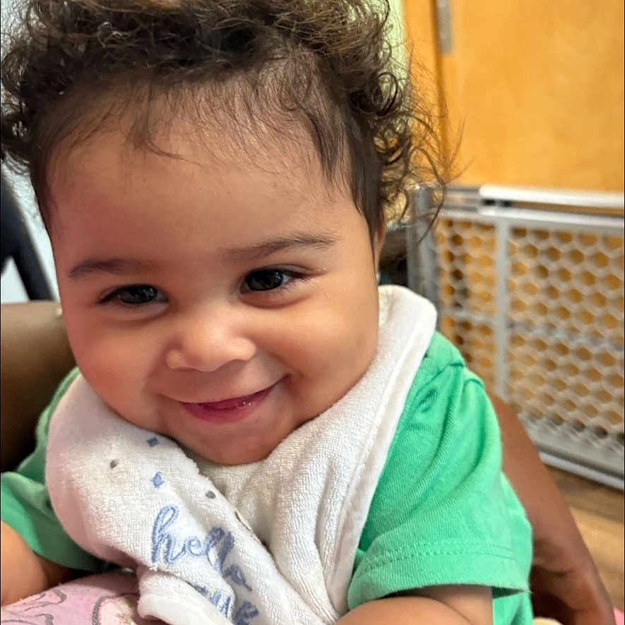Marciano joined the competition — help win amazing prizes! baby, child, smiling, curly_hair, green_shirt, bib, indoor, baby_gate, wooden_door, happy, face, portrait, cute, infant, person, skin, sitting, closeup, young_child, playful