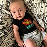 baby, black_shirt, blanket, blue_eyes, child, closeup, colorful_graphic, cute, family_photo, gray_shorts, indoors, infant, looking_at_camera, lying_down, natural_light, patterned_fabric, portrait, relaxed, seated, wide_eyed