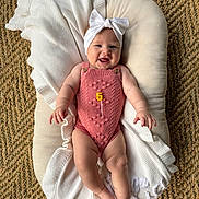 Lilia is registered to the contest to win money with this photo: baby, infant, smiling, headband, bow, pink_romper, knitted, blanket, cushion, woven_rug, floor, happy, cute, milestone, celebration, child, portrait, indoor, person, laying
