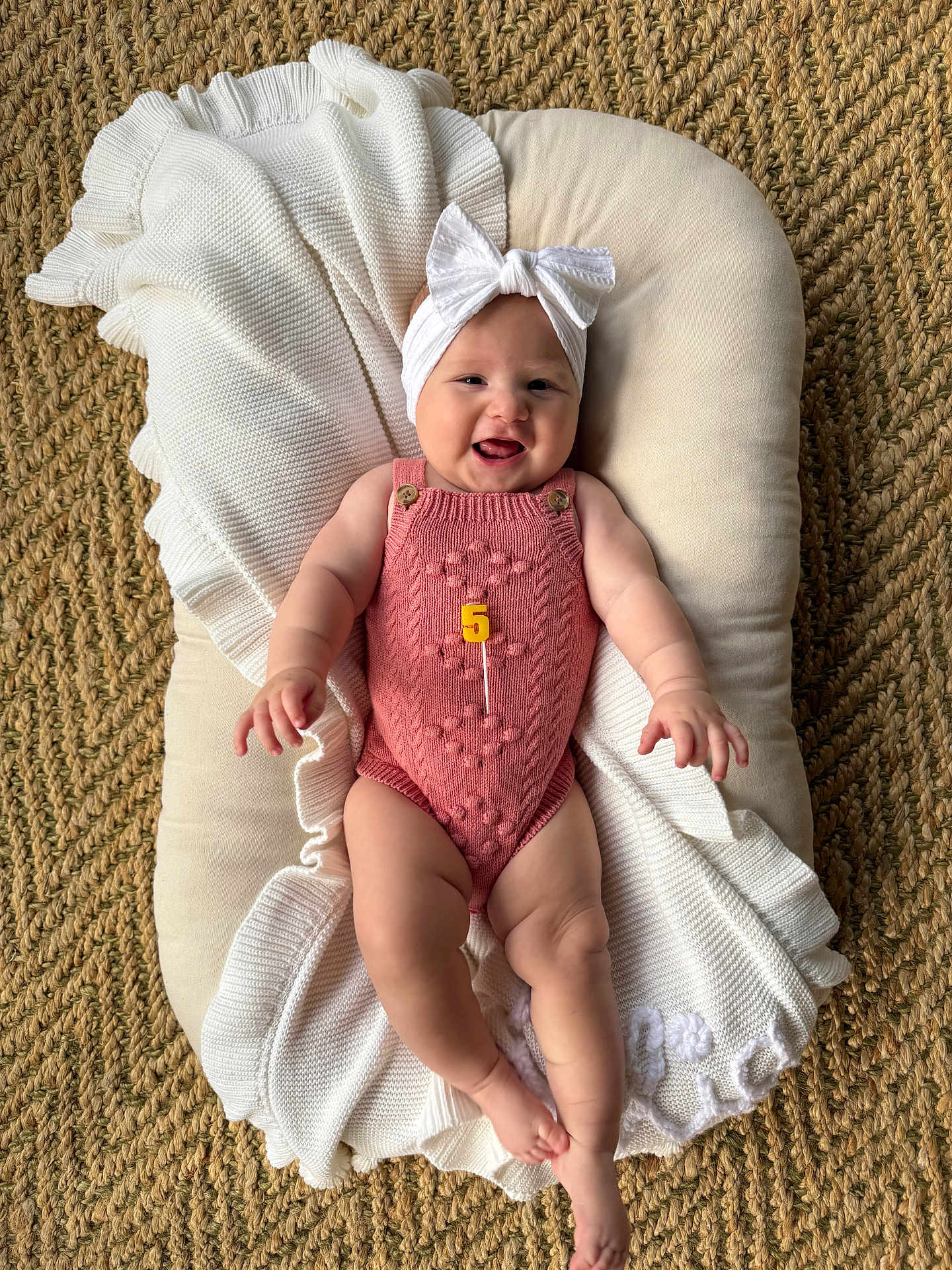 Lilia is registered to the contest to win money with this photo: baby, infant, smiling, headband, bow, pink_romper, knitted, blanket, cushion, woven_rug, floor, happy, cute, milestone, celebration, child, portrait, indoor, person, laying