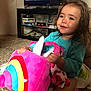 toddler, child, plush_toy, unicorn, rainbow, carpet, television, furniture, shelf, board_games, turtle, indoor, smiling, happy, clothing, person, hand, toy, hair, home
