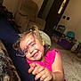 child, girl, smiling, glasses, pigtails, pink_clothing, pointing, indoor, carpet, armchair, toys, storage_bins, happy, person, portrait, casual, home, cute, young_child, playful
