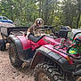 Lady is registered to the contest to win money with this photo: atv, bandana, beagle, budweiser, can, dirt_road, dog, forest, happy, nature, outdoor, pet, quad_bike, red_vehicle, rocks, sunlight, tire, trailer, trees, vehicle