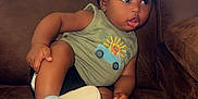 Camryn is registered to the contest to win money with this photo: toddler, child, baby, cap, green_tank_top, cartoon_print, black_shorts, blue_shoes, couch, brown_couch, indoor, furniture, cute, curious, expression, sitting, person, face, headwear, casual_clothing