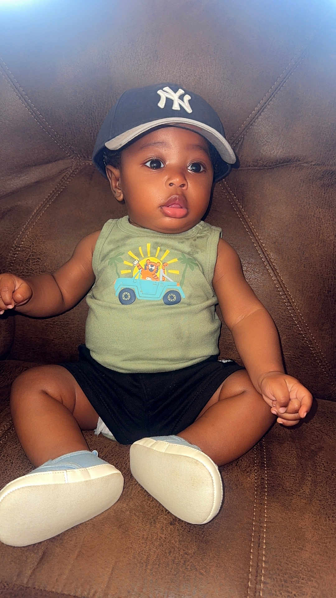 Camryn is registered to the contest to win money with this photo: baby, child, sitting, cap, hat, tank_top, shorts, shoes, leather_chair, curious, wide_eyes, indoor, cute, infant, person, clothing, footwear, portrait, expression, young