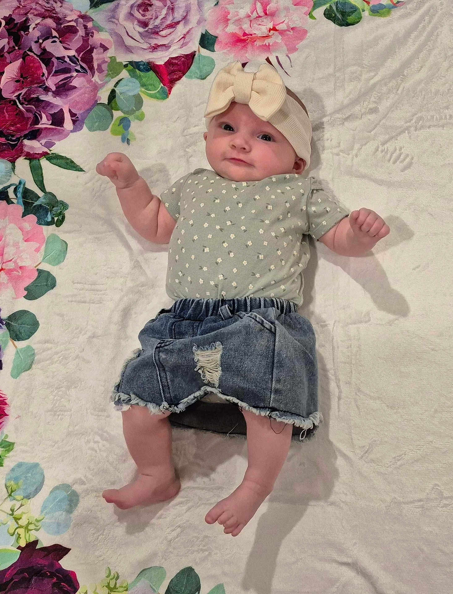 Violet is registered to the contest to win money with this photo: baby, infant, headband, bow, denim_shorts, floral_blanket, patterned_clothing, cute, child, lying_down, pink_flowers, green_leaves, soft_texture, skin, barefoot, small_hands, small_feet, portrait, indoors, happy_expression