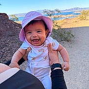 Janelle is registered to the contest to win money with this photo: baby, child, smiling, hat, purple_hat, outdoor, nature, lake, mountains, rocks, desert, person, hand, holding, happy, sunny, toddler, clothing, short_sleeves, daylight