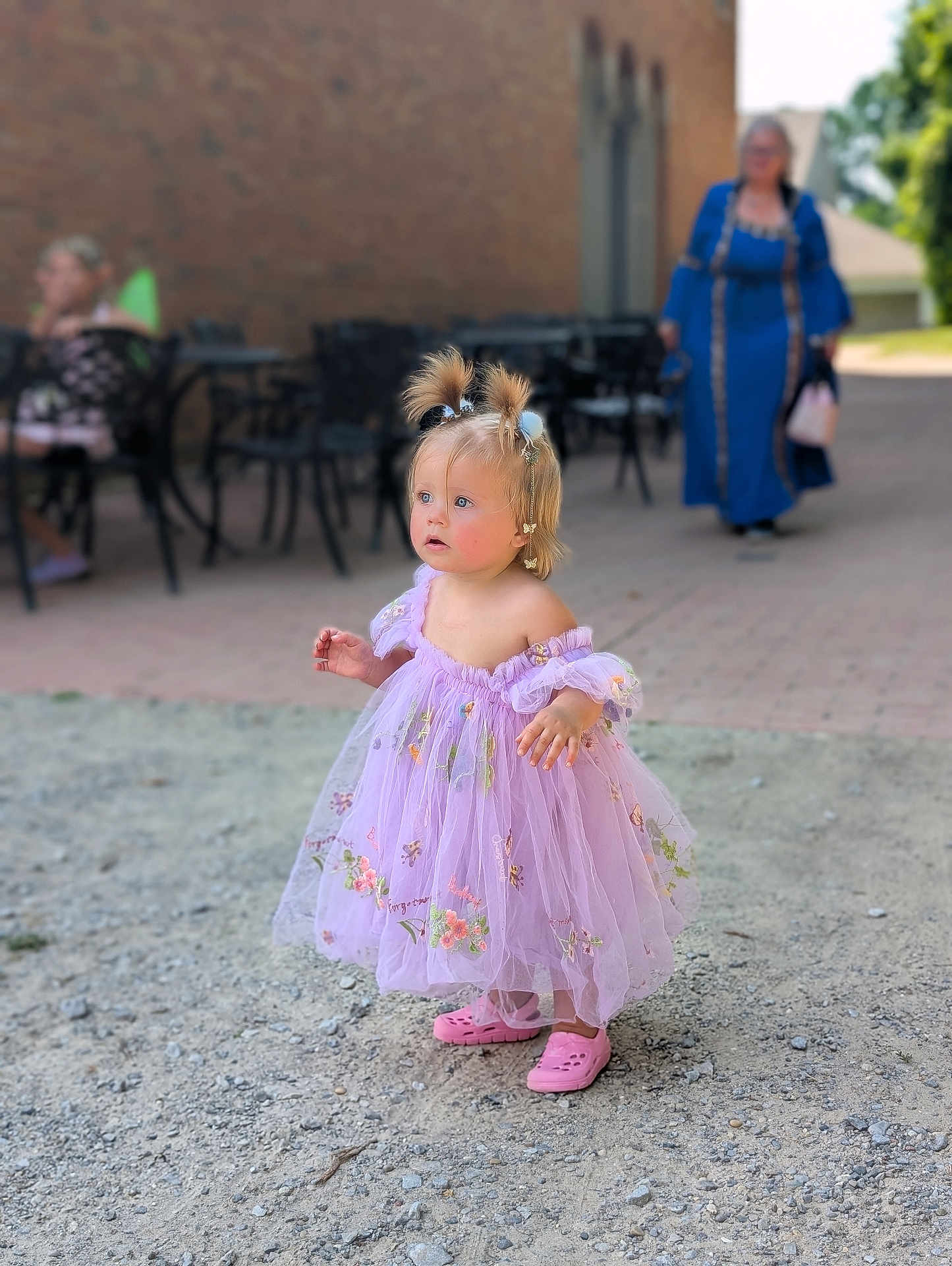 Ridlee Renee is registered to the contest to win money with this photo: child, toddler, dress, lavender, pink_shoes, pigtails, hair_accessories, outdoor, gravel, curious_expression, person, blurred_background, adult, chairs, table, sidewalk, sunny, daytime, candid, portrait
