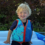 Soren is registered to the contest to win money with this photo: child, toddler, smiling, life_vest, inflatable_pool, outdoor, summer, blonde_hair, happy, water, blue, greenery, nature, play, sunlight, casual_clothing, person, recreation, fun, vacation