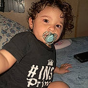 Alexis is registered to the contest to win money with this photo: toddler, child, curly_hair, pacifier, black_shirt, indoor, bed, pillow, baby_lotion, hand, face, cute, infant, person, young_child, casual, expression, closeup, skin, sitting
