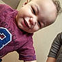baby, child, smiling, teeth, closeup, portrait, indoor, tshirt, maroon_shirt, arm, adult_arm, candid, playful, curly_hair, forehead, wall, hardwood_floor, person, happy, squinting