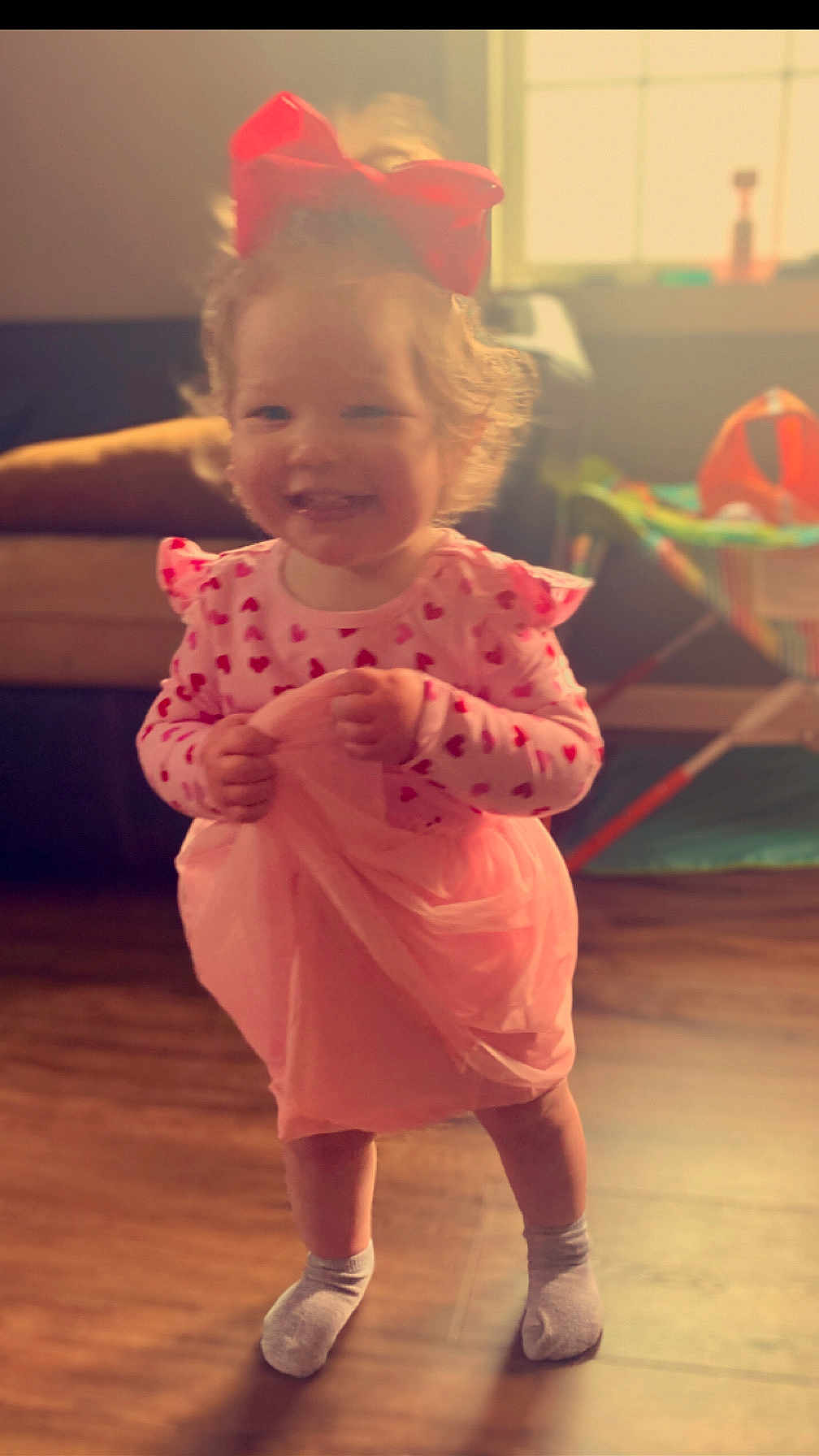 Lorelei is registered to the contest to win money with this photo: toddler, child, indoor, smiling, dress, bow, pink, hearts, socks, wooden_floor, window, sunlight, play_area, happy, person, curly_hair, cute, standing, fabric, home