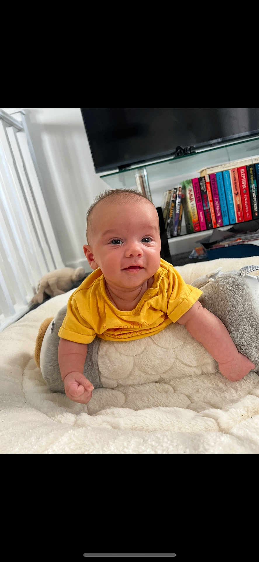 Carson joined the competition — help win amazing prizes! baby, infant, yellow_shirt, smile, face, plush, cushion, indoor, bookshelf, books, television, soft_toys, blanket, person, home, child, curious, happy, floor, furniture