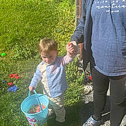 Greyson is registered to the contest to win money with this photo: adult, bucket, casual, child, clothing, daylight, footwear, garden, grass, greenery, hand_holding, holding, outdoor, pants, person, play, shirt, sidewalk, sunlight, toy