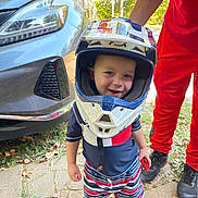 Greyson is registered to the contest to win money with this photo: toddler, child, helmet, motocross_helmet, toy_car, car, adult, red_clothing, blue_clothing, outdoor, sidewalk, grass, leaves, smiling, happy, footwear, person, vehicle, daylight, portrait