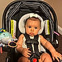 baby, bow, car_seat, car_seat_harness, carpet, child, clothing, curious_expression, cute, face, head, indoor, infant, person, plush_toy, safety, sitting, soft_lighting, straps, toy