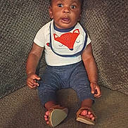 Nolan is registered to the contest to win money with this photo: baby, child, infant, sitting, couch, bib, fox_design, sandals, clothing, face, eyes, expression, indoors, texture, footwear, person, young_child, cute, looking, portrait