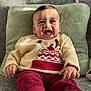 baby, child, smiling, sitting, sweater, red_pants, pom_pom, couch, cushion, indoor, happy, cute, face, portrait, hands, clothing, cozy, infant, person, warm
