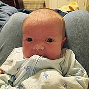 Scarlett is registered to the contest to win money with this photo: baby, infant, clothing, onesie, shark_pattern, chewing, face, head, eyes, person, lap, jeans, indoor, cute, young_child, newborn, skin, hand, sleeve, cozy