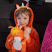Cain-Leigh joined the competition — help win amazing prizes! child, costume, orange, plush_toy, dragon, couch, smiling, indoor, clothing, toy, person, face, hood, fabric, blanket, folded_clothes, cute, sitting, happy, cozy