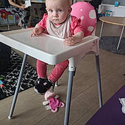 Bethany is registered to the contest to win money with this photo: baby, high_chair, pink_clothing, indoor, child, woman, reading, book, floor, toy, carpet, table, chair, person, footwear, furniture, room, expression, blonde_hair, polka_dot