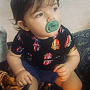 Ezra is registered to the contest to win money with this photo: toddler, child, pacifier, shirt, shorts, sitting, indoor, blanket, patterned, curly_hair, face, looking_away, hands, legs, comfort, expression, colorful_clothing, young_child, portrait, casual