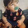 toddler, child, pacifier, shirt, shorts, sitting, indoor, blanket, patterned, curly_hair, face, looking_away, hands, legs, comfort, expression, colorful_clothing, young_child, portrait, casual