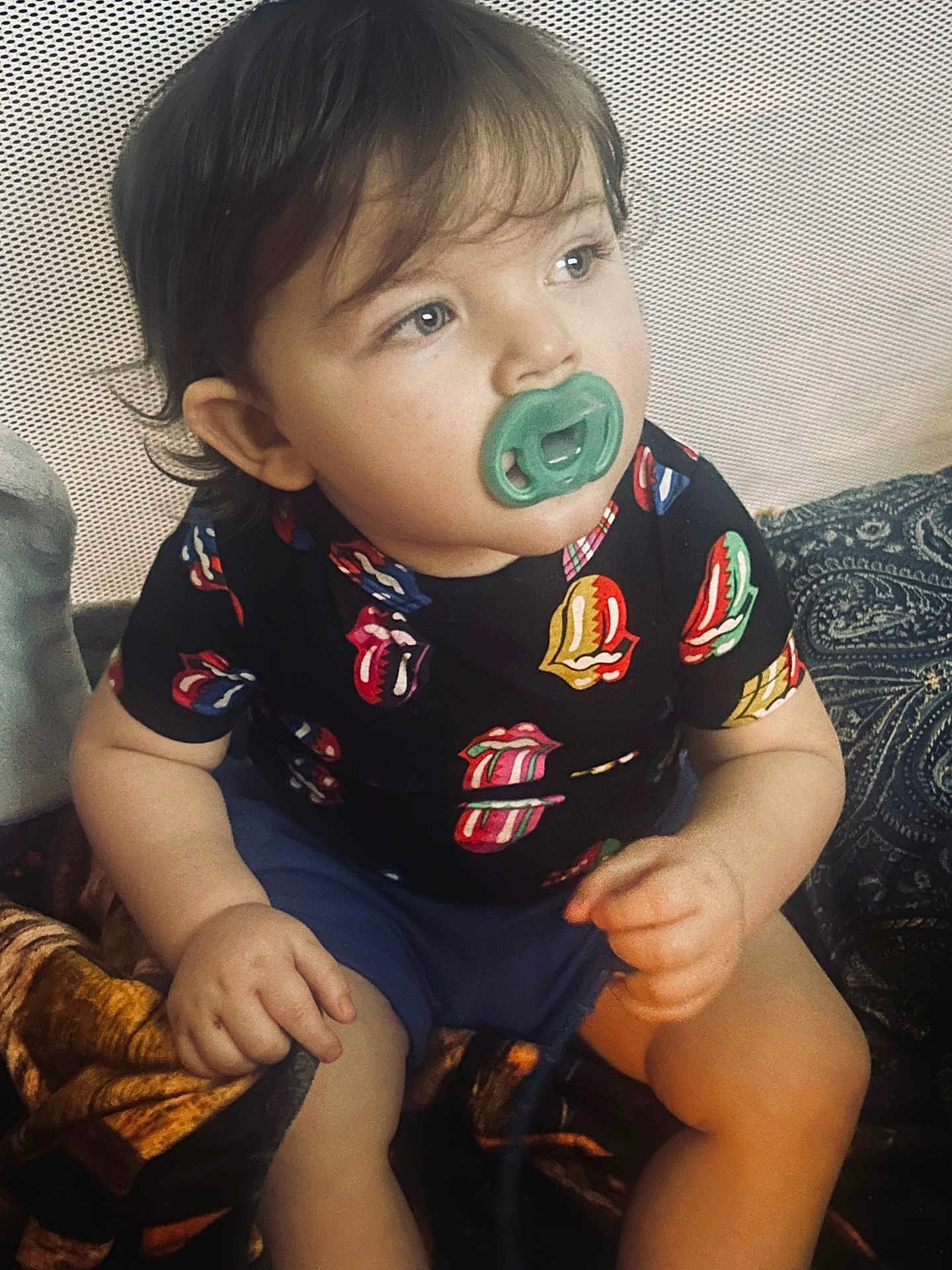 Ezra is registered to the contest to win money with this photo: toddler, child, pacifier, shirt, shorts, sitting, indoor, blanket, patterned, curly_hair, face, looking_away, hands, legs, comfort, expression, colorful_clothing, young_child, portrait, casual