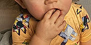 Ezra is registered to the contest to win money with this photo: child, toddler, finger_in_mouth, yellow_shirt, cartoon_print, curly_hair, blue_eyes, indoor, seated, soft_chair, skin, short_sleeves, cute, closeup, portrait, expression, casual, young_child, comfort, colorful_clothing
