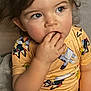 child, toddler, finger_in_mouth, yellow_shirt, cartoon_print, curly_hair, blue_eyes, indoor, seated, soft_chair, skin, short_sleeves, cute, closeup, portrait, expression, casual, young_child, comfort, colorful_clothing