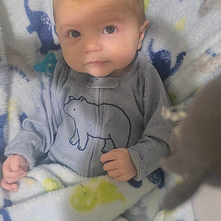 Theon is registered to the contest to win money with this photo: baby, blanket, onesie, bear_design, dinosaur_pattern, infant, cute, face, portrait, clothing, soft_texture, blue, yellow, grey, child, indoor, lying_down, young, newborn, comfort