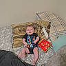 Theon is registered to the contest to win money with this photo: baby, bed, pillow, blanket, onesie, toy, book, indoor, child, infant, face, clothing, fabric, pattern, person, resting, lying_down, soft, comfortable, colorful