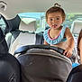 toddler, child, car, car_seat, interior, tongue_out, playful, person, seat, headrest, window, daylight, casual_clothing, hair_tie, backseat, vehicle, young_child, cute, portrait, funny_expression