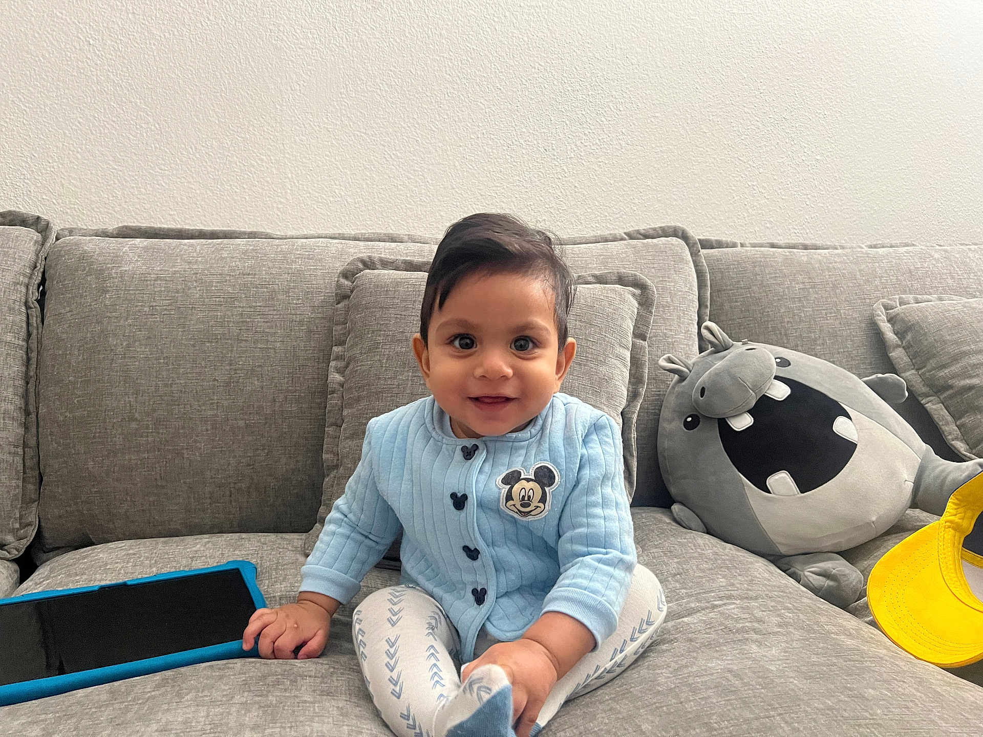 Ezhaan is registered to the contest to win money with this photo: baby, child, smiling, blue_sweater, mickey_mouse, couch, gray_cushions, plush_hippo, yellow_cap, tablet, indoor, happy, sitting, person, face, cute, home, cozy, soft_lighting, casual